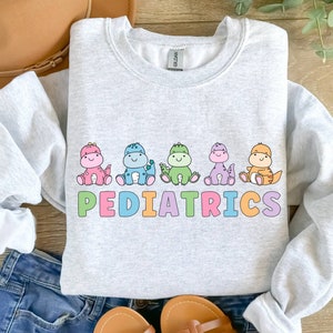 Peut inclure: Un sweat-shirt gris clair avec un motif de cinq dinosaures color&eacute;s et le mot "PEDIATRICS" en lettres arc-en-ciel.