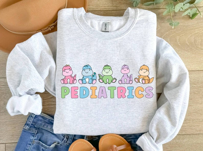 Peut inclure: Un sweat-shirt gris clair avec un motif de cinq dinosaures color&eacute;s et le mot "PEDIATRICS" en lettres arc-en-ciel.