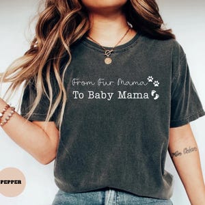 May include: Dark grey t-shirt with the text "From Fur Mama To Baby Mama" in white script. The shirt features paw prints and baby footprints. The shirt is a comfortable, casual style.