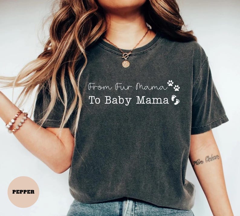 May include: Dark grey t-shirt with the text "From Fur Mama To Baby Mama" in white script. The shirt features paw prints and baby footprints. The shirt is a comfortable, casual style.