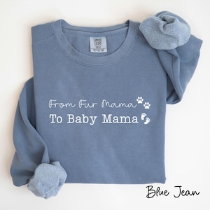 May include: Blue jean colored sweatshirt with the text "From Fur Mama To Baby Mama" in white script. The design includes paw prints and baby footprints. The sweatshirt has long sleeves and a crew neck.
