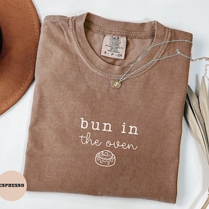 May include: A brown short-sleeved t-shirt with a crew neckline and a Comfort Colors label. The front features the white text "bun in the oven" above a line drawing of a cinnamon roll. A silver-toned necklace rests on the fabric.