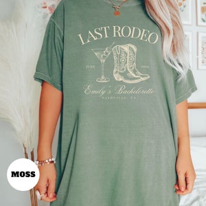 May include: A green t-shirt with the text "LAST RODEO" printed on the front. The text is in white and is surrounded by a drawing of a pair of cowboy boots and a martini glass. The text "JUNE 2024" is printed below the boots and the text "Emily's Bachelorette NASHVILLE, TN" is printed below the martini glass.