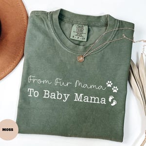 May include: An olive green t-shirt with the text "From Fur Mama To Baby Mama" in white script, along with paw prints and baby footprints. A gold necklace with a pendant rests on the shirt. The shirt is labeled "Comfort Colors".