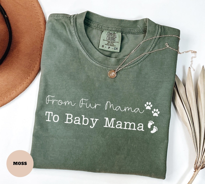 May include: An olive green t-shirt with the text "From Fur Mama To Baby Mama" in white script, along with paw prints and baby footprints. A gold necklace with a pendant rests on the shirt. The shirt is labeled "Comfort Colors".
