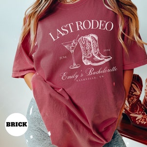 May include: A maroon t-shirt with the text "Last Rodeo" in white, featuring a pair of cowboy boots and a martini glass. The text "Emily's Bachelorette" is also in white, along with the date "June 2024" and the location "Nashville, TN".
