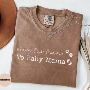 May include: Espresso-colored t-shirt with the text "From Fur Mama To Baby Mama" in white script. The shirt features paw prints and baby footprints. A silver necklace with a pendant rests on the shirt.
