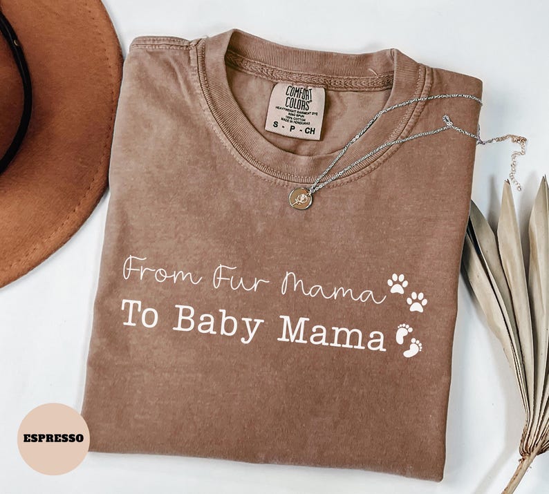 May include: Espresso-colored t-shirt with the text "From Fur Mama To Baby Mama" in white script. The shirt features paw prints and baby footprints. A silver necklace with a pendant rests on the shirt.