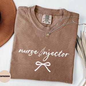 May include: Espresso-colored t-shirt with the words "nurse injector" and a syringe graphic in white. The shirt has a crew neck and a small tag at the neckline. A silver necklace with a pendant is draped across the shirt.