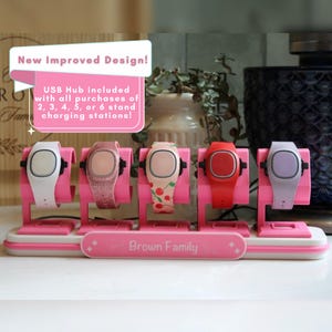 May include: A pink charging station stand holding five smartwatches with various bands, including white, glittery pink, cherry-patterned, red, and light purple. The base features the text Brown Family and a sign stating New Improved Design.