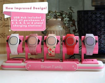 Op de afbeelding: Een roze oplaadstation met vijf smartwatches met diverse bandjes, waaronder wit, glitterroze, kersenpatroon, rood en lichtpaars. De basis bevat de tekst Brown Family en een bordje met de tekst New Improved Design.