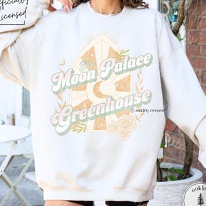 Op de afbeelding: Crèmekleurige sweatshirt met de tekst "The Moon Palace Greenhouse" in een retro lettertype, met een zon, maan en bloemenontwerp. De sweatshirt heeft lange mouwen en een relaxte pasvorm. De woorden "Officially Licensed" staan linksboven.