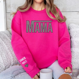 Op de afbeelding: Roze sweatshirt met een zwart-wit geblokte "MAMA" grafiek op de voorkant. De linkermouw heeft een zwart-wit geblokt hart met de tekst "Xander Violet" eronder.