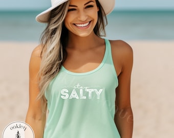 May include: A woman wearing a mint green tank top with the text "stay salty" in white. She is wearing a white sun hat and is standing on a beach.