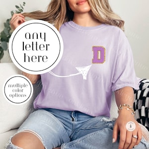 May include: A light purple t-shirt with a white arrow pointing to a gold and purple letter D. The text "any letter here" is written in a circle above the arrow. The text "multiple color options" is written in a circle below the arrow.