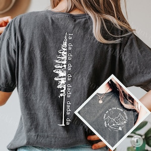 Könnte beinhalten: Ein dunkelgraues T-Shirt mit einer weißen Silhouette eines Kiefernbaums und dem Text "la da da da da dada dada da" auf der Rückseite. Die Vorderseite des Shirts zeigt eine weiße Silhouette von Bergen und den Text "meet me in the woods tonight".