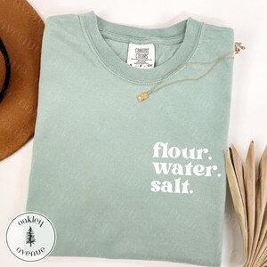 Op de afbeelding: Een saliegroen t-shirt met de woorden "flour. water. salt." in het wit. Een gouden ketting met een kleine hanger ligt op het gevouwen shirt. Het shirt heeft een Comfort Colors label.