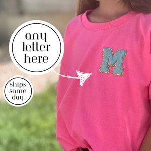 May include: Pink t-shirt with a blue felt letter M with gold glitter accents. The letter is sewn onto the shirt. The text "any letter here" is written on a white circle.