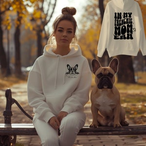 Op de afbeelding: Een witte hoodie met een Franse Bulldog-afbeelding en de tekst "Amelie's Mom" wordt gedragen door een persoon. Een tweede hoodie met de tekst "IN MY FRENCHIE MOM ERA" wordt getoond. Een bruine Franse Bulldog zit ernaast.