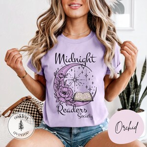 以下が含まれることがあります： 「Midnight Readers Society」の文字と三日月デザインが特徴の薄紫色のTシャツ。デザインには花、星、開いた本が含まれています。このTシャツは柔らかく快適な素材で作られています。