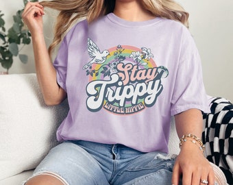 May include: A lavender t-shirt with a retro graphic design featuring a white bird, flowers, and a rainbow. The text on the shirt reads "Stay Trippy Little Hippie".