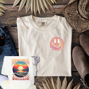 以下が含まれることがあります： グラフィックデザインが施されたクリーム色のTシャツ2枚。1枚は「FOREVER CHASING SUNSETS」という文字と夕焼けが描かれている。もう1枚は夕焼けを背景にスマイリーフェイスが描かれている。