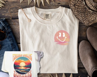 May include: Two cream colored t-shirts with graphic designs. One shirt features a sunset with the text "FOREVER CHASING SUNSETS". The other shirt features a smiley face with a sunset background.