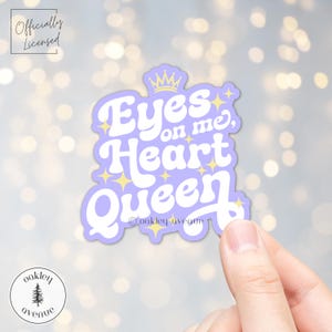 Könnte beinhalten: Ein hellvioletter Aufkleber mit dem Text "Eyes on me, Heart Queen" in Weiß, verziert mit einer goldenen Krone und Sternen. Der Aufkleber wird von einer Hand vor einem verschwommenen Hintergrund gehalten. Der Aufkleber hat auch den Text "oakley avenue" und "Officially Licensed".