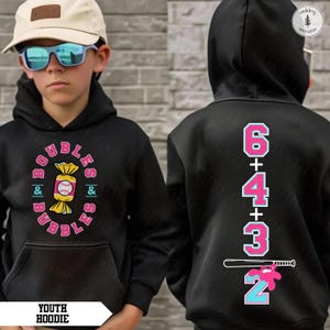 May include: Black youth hoodie with a front graphic of a baseball inside a candy wrapper, surrounded by the words "Doubles & Bubbles". The back features the numbers 6, 4, 3, and 2 with a baseball bat.