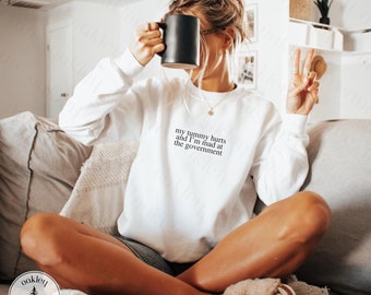 May include: White sweatshirt with the text "my tummy hurts and I'm mad at the government" printed on the front.