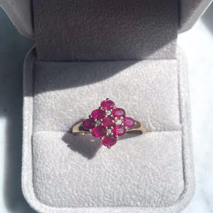 Pode incluir: Um anel de ouro com um conjunto de pedras preciosas vermelhas escuras de corte oval, realçado com pequenos diamantes transparentes. O anel é cravejado em uma banda de ouro e apresentado em uma caixa de joias de veludo cinza.
