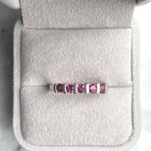 May include: A silver ring with five vibrant pink gemstones set in a row. The ring is displayed inside a gray velvet jewelry box, showcasing the gemstones' color and the ring's design. The gemstones are evenly spaced.