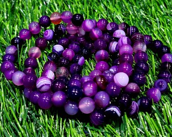 May include: A close-up of a purple and white agate gemstone bracelet. The bracelet is made of multiple strands of beads, each with a different pattern of purple and white stripes.
