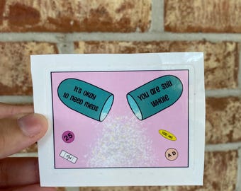 May include: A sticker with a pink background and two teal pill-shaped capsules. The left capsule says "It's okay to need meds" and the right capsule says "You are still whole". There are two smaller pills, one pink and one yellow, with text on them.