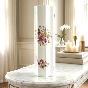 May include: A tall, white, hexagonal ceramic vase featuring delicate, colorful floral bouquets painted on its sides. The vase has a smooth, glossy finish and a straight, vertical structure, showcasing detailed illustrations of pink, yellow, and blue flowers.