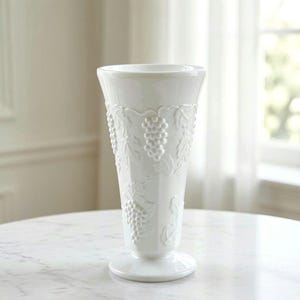 May include: A white ceramic vase with a flared rim and a pedestal base. The exterior surface features a raised, embossed pattern of grape clusters and vine leaves that wrap around the body of the vessel, creating a textured, monochromatic design.