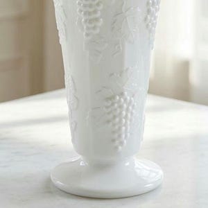 May include: A white glass vase featuring a raised, embossed pattern of grape clusters and vine leaves. The vase has a tapered cylindrical body that widens towards the top and sits on a circular pedestal base with a smooth, glossy finish.