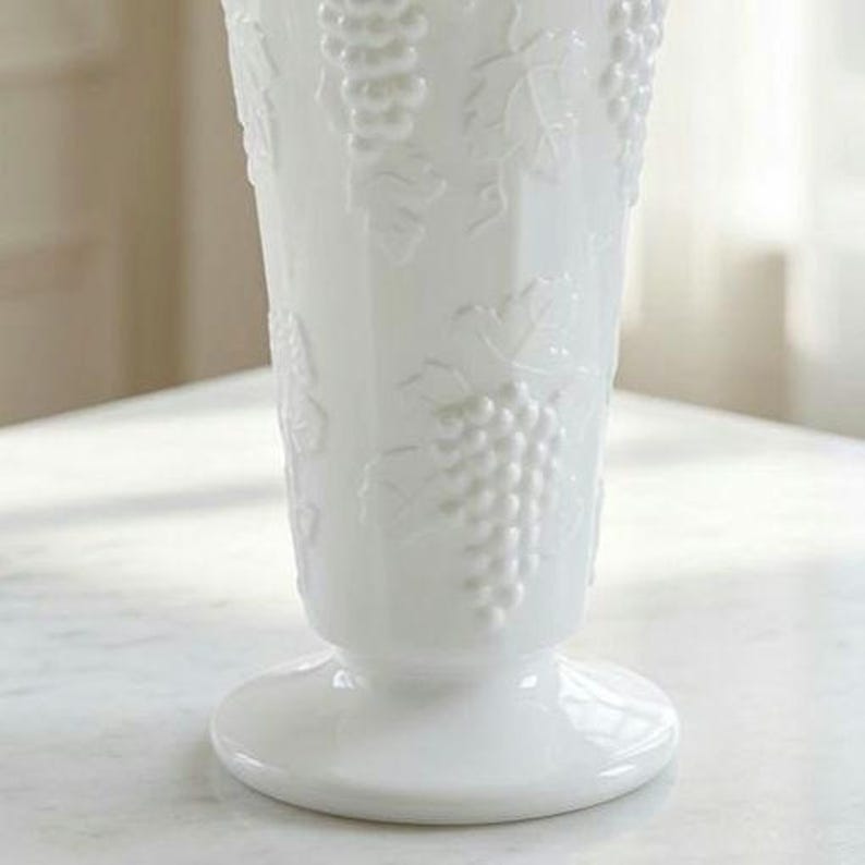 May include: A white glass vase featuring a raised, embossed pattern of grape clusters and vine leaves. The vase has a tapered cylindrical body that widens towards the top and sits on a circular pedestal base with a smooth, glossy finish.