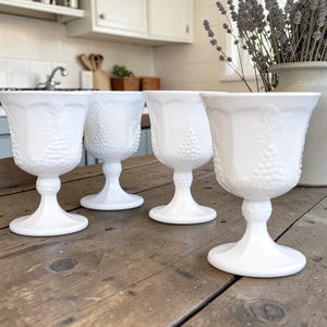 May include: A set of four white milk glass goblets featuring a raised relief pattern of grape clusters and leaves on the bowl and a textured, beaded design on the stem. Each glass has a wide, circular base and a smooth, flared rim for drinking.