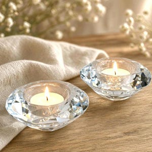 May include: Two clear, faceted glass tea light candle holders with a diamond-like shape. Each holder contains a small, lit white candle with a visible flame, casting a warm glow through the transparent, geometric glass surfaces.