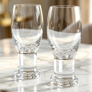 May include: Two clear glass tumblers with a rounded bowl shape that tapers toward a thick, cylindrical base. The glasses feature a smooth, transparent surface and a sturdy, weighted bottom design, standing upright with a polished, reflective finish.