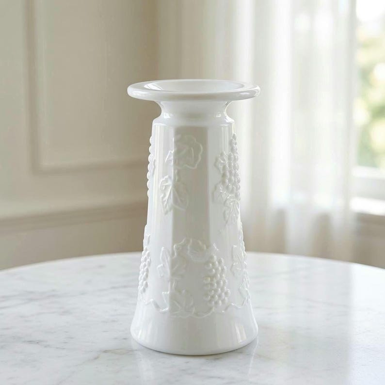 May include: A white ceramic vase with a flared rim and a tapered body featuring a raised, embossed pattern of grape clusters and vine leaves wrapping around the exterior. The surface has a smooth, glossy finish and a solid white colour throughout.