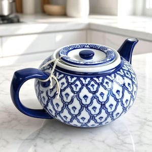 May include: A round ceramic teapot featuring a white base with a repeating blue scalloped pattern. The teapot has a solid blue handle, a matching blue spout, and a circular lid with a blue knob. A thin white string is tied around the handle.