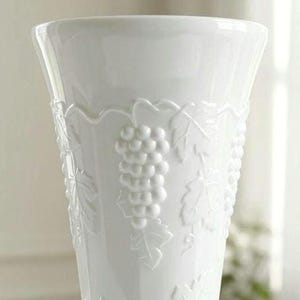 May include: A white ceramic vase featuring a raised, embossed pattern of grape clusters and vine leaves encircling the exterior. The vessel has a wide, flared rim and a smooth, glossy finish that highlights the intricate, monochromatic relief design.