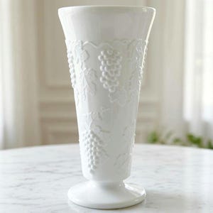 May include: A tall, white glass vase featuring a raised, embossed pattern of grape clusters and vine leaves. The vase has a flared rim, a tapered body, and a circular pedestal base, all finished in a smooth, glossy, opaque white material.