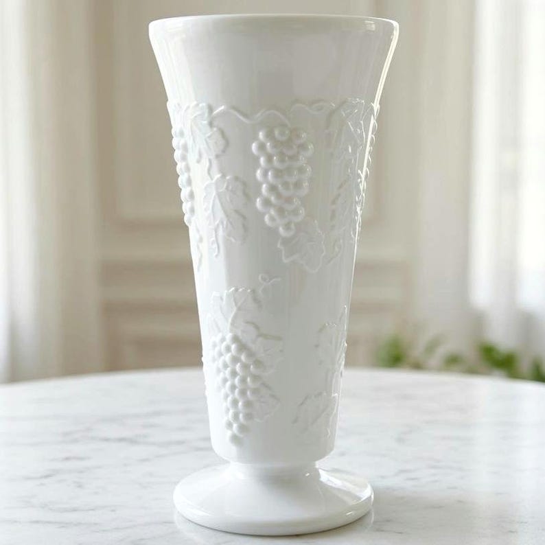 May include: A tall, white glass vase featuring a raised, embossed pattern of grape clusters and vine leaves. The vase has a flared rim, a tapered body, and a circular pedestal base, all finished in a smooth, glossy, opaque white material.