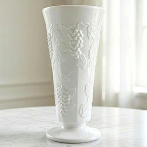 May include: A tall, white ceramic vase featuring a raised, embossed pattern of grape clusters and vine leaves wrapping around the exterior. The vase has a flared rim, a tapered body, and a circular pedestal base with a smooth, glossy finish.