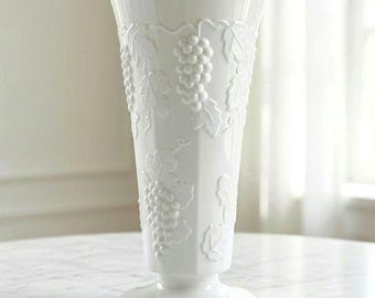 May include: A tall, white ceramic vase featuring a raised, embossed pattern of grape clusters and vine leaves wrapping around the exterior. The vase has a flared rim, a tapered body, and a circular pedestal base with a smooth, glossy finish.