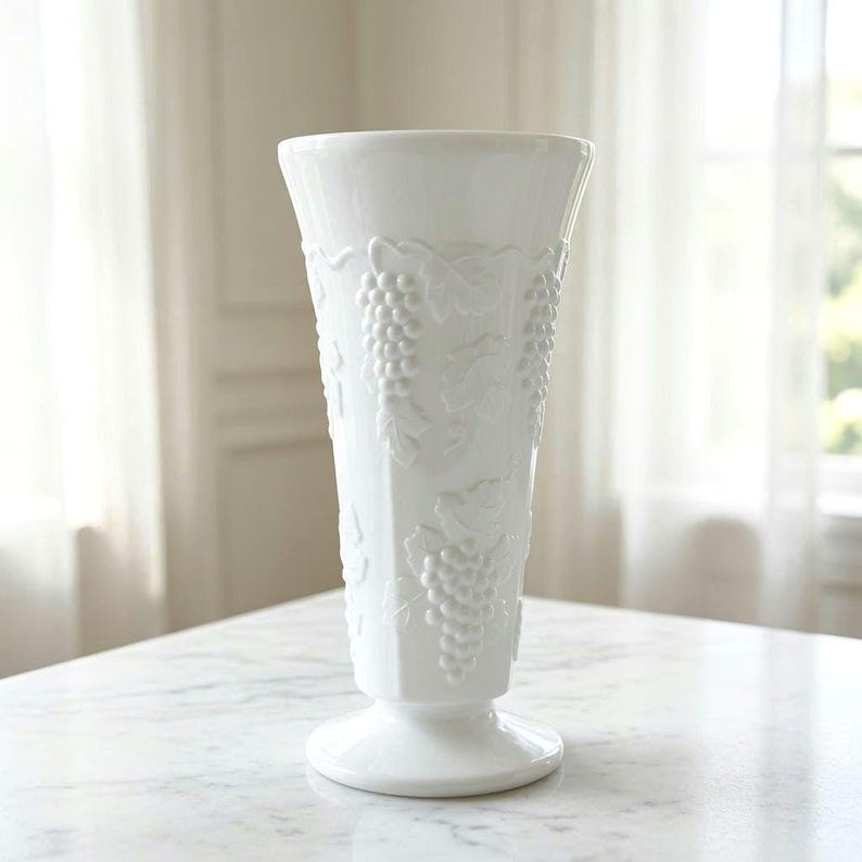 May include: A tall, white milk glass vase featuring a flared rim and a pedestal base. The exterior surface is decorated with a raised, embossed pattern of grape clusters and vine leaves that wrap around the body of the vessel in a repeating design.