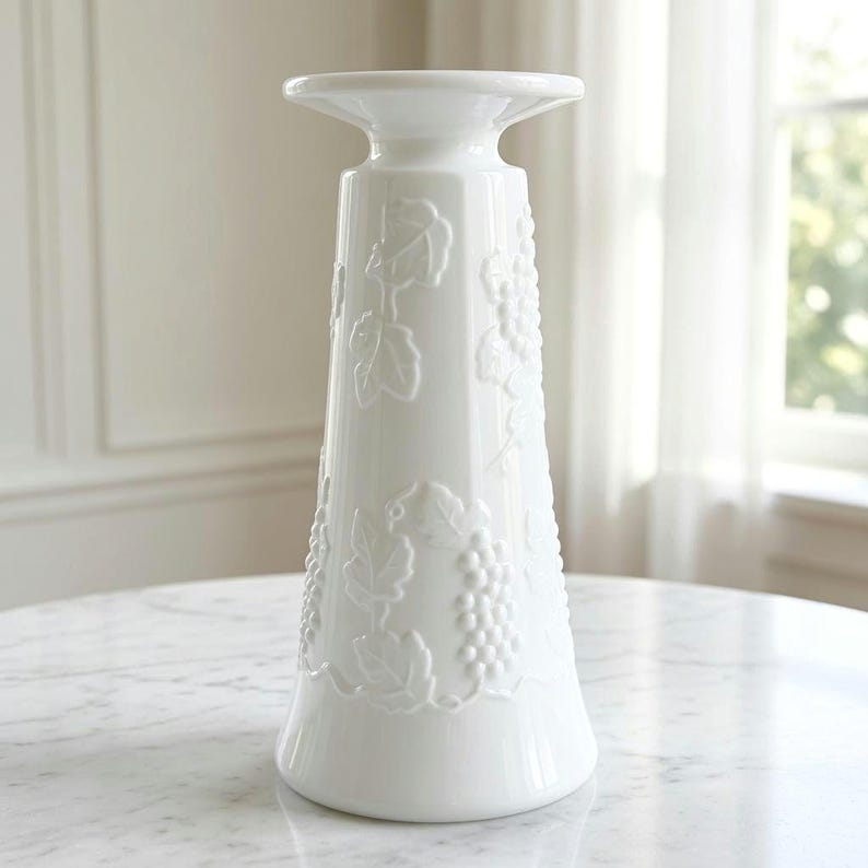 May include: A white ceramic pillar candle holder featuring a raised, embossed design of grape clusters and vine leaves wrapping around the tapered body. The piece has a wide, flat top platform and a flared base with a glossy finish.